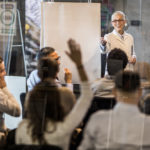 Happy senior doctor talking to large group of people on a seminar in a board room while aiming at one of them who wants to ask something. The view is through glass.