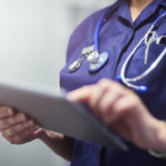 Female surgeon typing on digital tablet in hospital or surgery. She is wearing a dark blue nurse’s top and has her stethoscope around her neck. She is looking at her patients records on her digital tablet and sending emails.