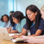 A multi-ethnic group of female nursing students are attending class together. They are seated at a long table in a classroom. The individuals are working in pairs on a project. The two individuals seated closest to the camera are having a discussion while using a laptop computer.