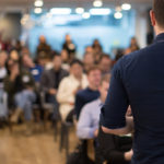 Conference photo audience and speaker giving speech. Seminar presenter on a panel during forum. Corporate manager in sales executive training discussion on stage. Investor pitch presentation. Speaker addressing group. Businessman entrepreneur giving a lecture to a sold-out crowd in a lecture hall. Speaker talking in seminar. Speaker at business conference or presentation stock photo. Male CEO talking during a business presentation in a board room. Copy space. Business coach concept.