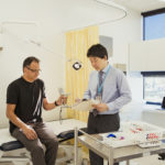 A front-view shot of a man using a hand grip strength test to test his maximum handgrip strength, a doctor can be seen looking at the results.