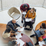 A professional team hold their regular meeting. They are a diverse group of multi-ethnicities and age ranges. They can be seen mid-discussion and are holding documents and laptops.