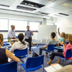 Two men standing in front of a team of people as they sit and listen to her giving a presentation