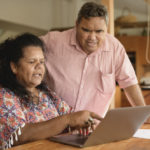Mature couple using laptop with woman pointing Aboriginal couple in their 50s at home using the internet, man leaning over woman and looking at screen