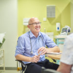 senior male gp at his desk chatting with a patient