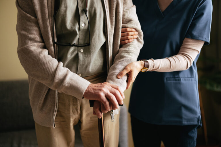 Unrecognizable Nurse Helping Senior Man To Walk In His Living Room An anonymous Japanese female caregiver supporting elderly patient to stand and walk with a walking cane at the nursing home.